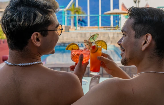Dos personas se miran y brindan con c&oacute;cteles junto a una piscina.