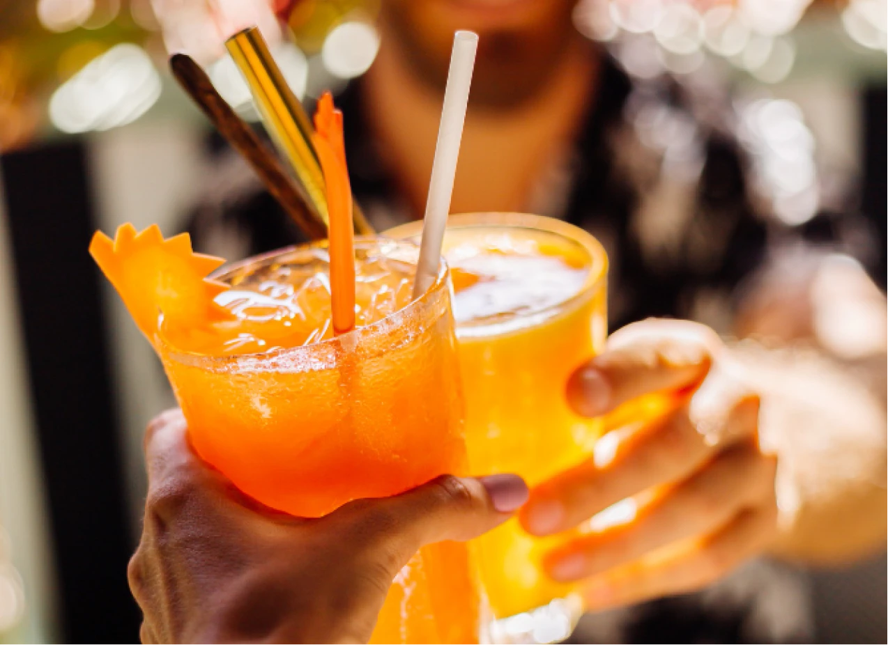 Manos brindan con c&oacute;ctel naranja con pajillas y cubo de hielo, fondo desenfocado