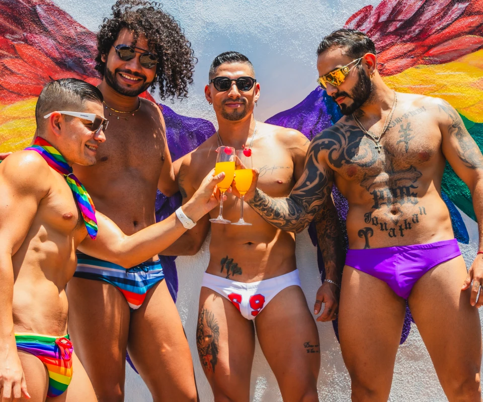 Cuatro hombres con trajes de ba&ntilde;o, gafas y bebidas en una fiesta colorida.