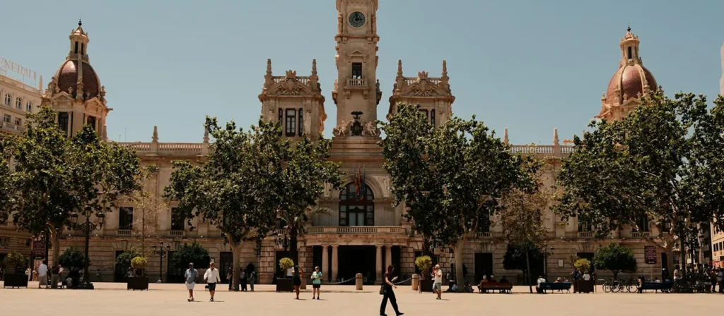 Edificio hist&oacute;rico con c&uacute;pulas y torres, &aacute;rboles y personas en una plaza