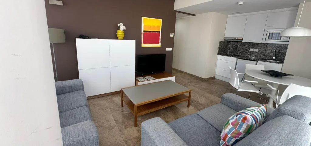 Sala de estar con sof&aacute;, mesa de centro y cocina blanca al fondo