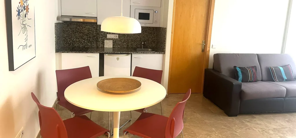 Cocina y comedor con mesa redonda blanca, sillas rojas y sof&aacute; gris junto a puerta