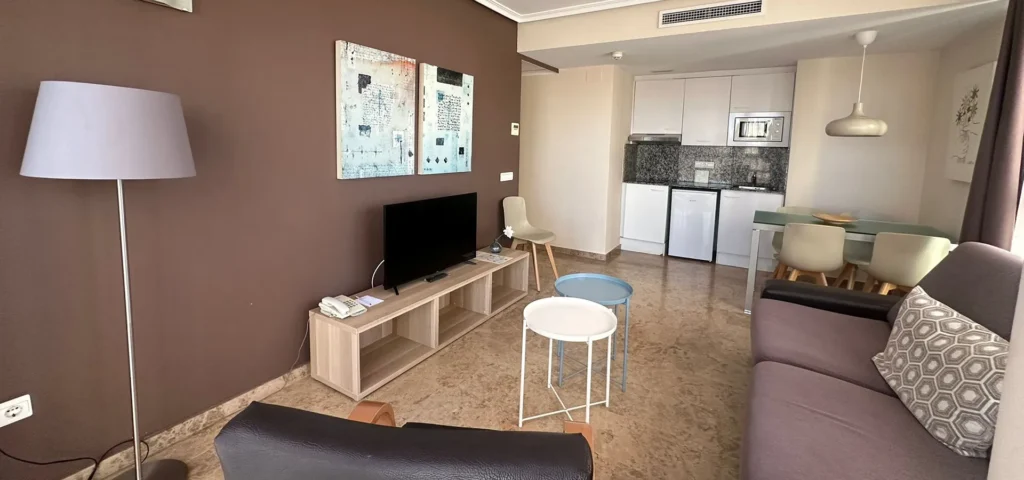 Sala de apartamento con sof&aacute;, TV, mesa y cocina con muebles blancos