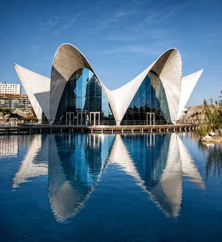 Edificio moderno con formas curvas reflejadas en un estanque bajo cielo azul.