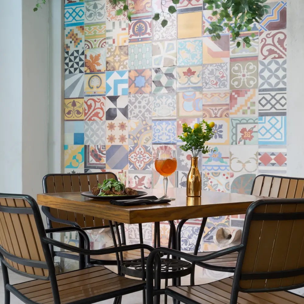 Mesa con dos sillas y copa con bebida frente a pared de azulejos coloridos