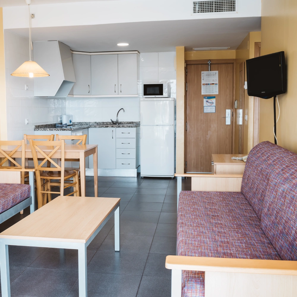 Cocina-comedor con sof&aacute;, mesa, muebles blancos y puerta de acceso.