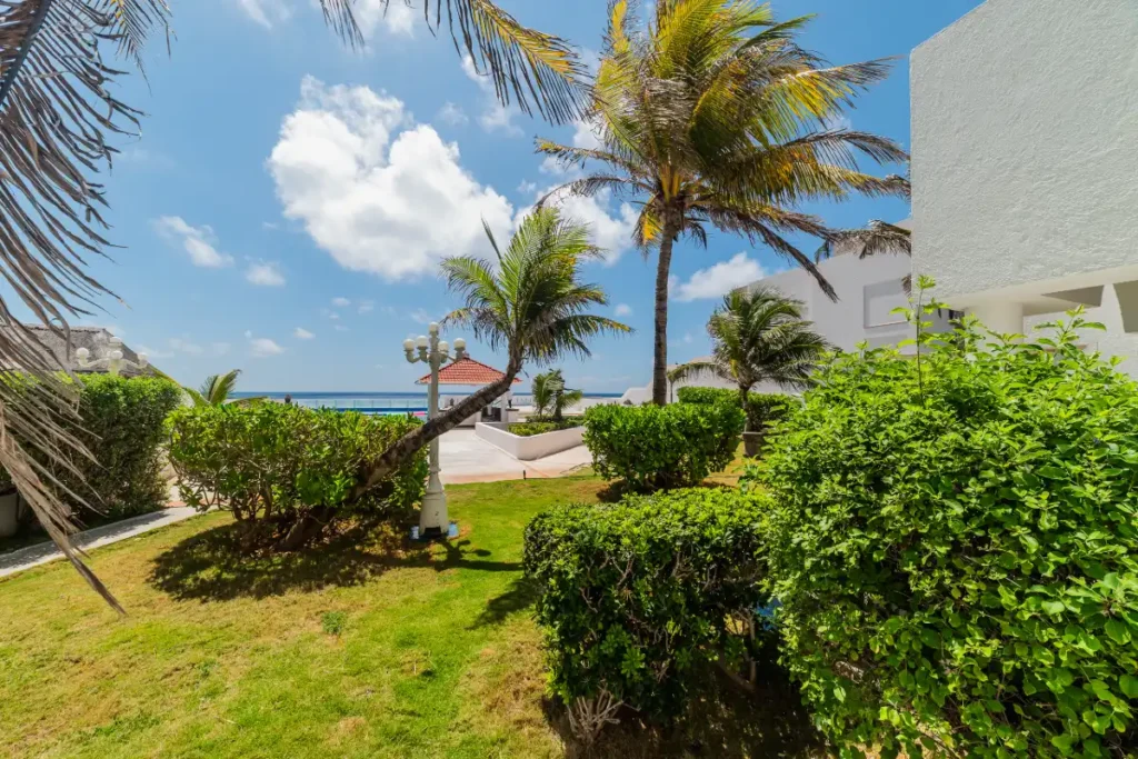 Jard&iacute;n tropical con palmeras, c&eacute;sped verde y vista al mar bajo cielos nublados.