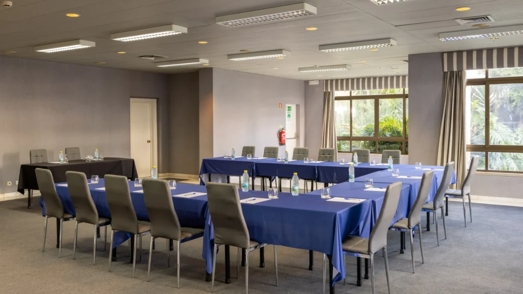 Sala de reuni&atilde;o com mesas retangulares e cadeiras, ilumina&ccedil;&atilde;o do teto e janelas.