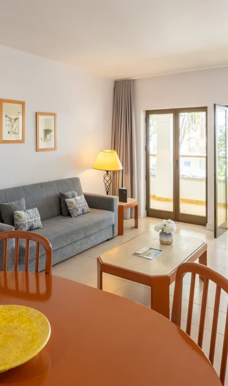 Sala de estar com sof&aacute; cinza, mesa de centro e janelas amplas com luz solar