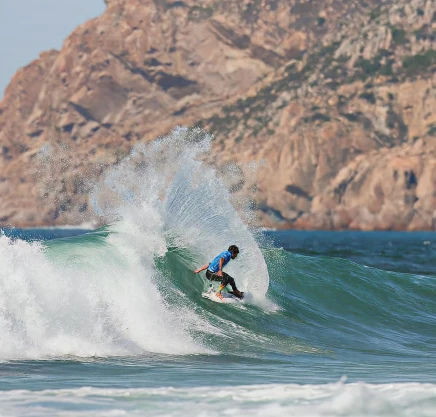 Surfista em p&eacute; numa onda, com montanhas ao fundo.