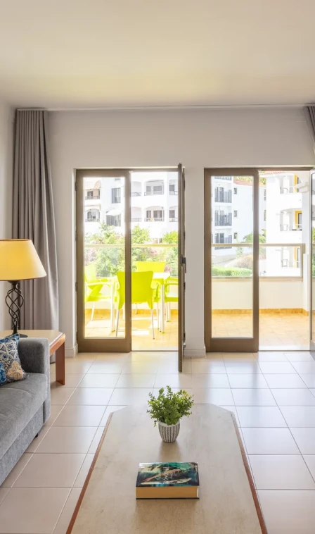 Sala com sof&aacute;, mesa e portas de vidro abertas para varanda ensolarada