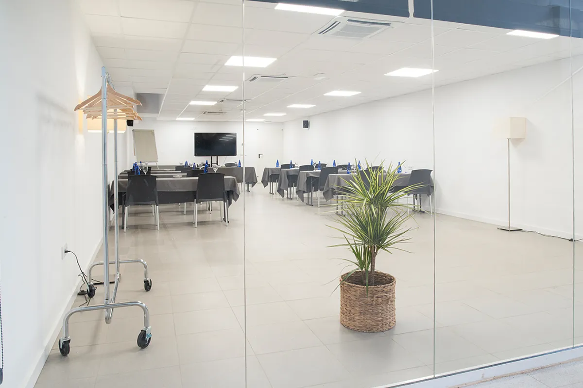 Sala de reuniones moderna con mesas, sillas y una planta junto a la entrada.