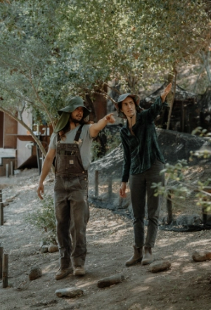 Two men in rugged clothing walk through a smoky outdoor settlement.
