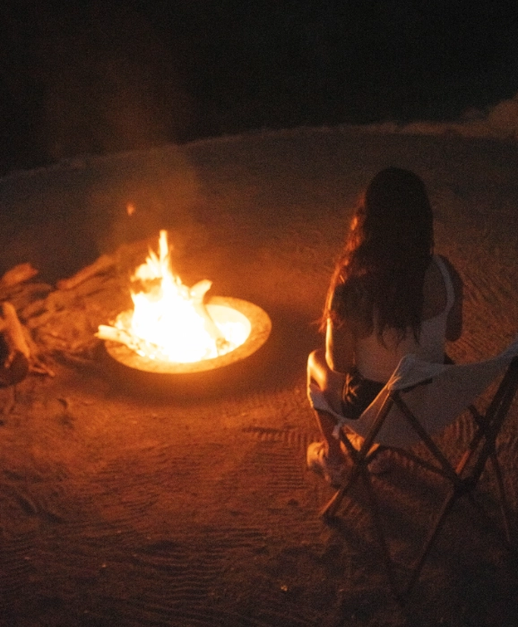 Woman kneels beside a campfire on a beach at night