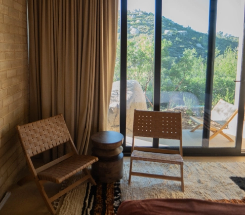 Balcony corner with wooden chairs, curtain, and view of green hills through glass doors