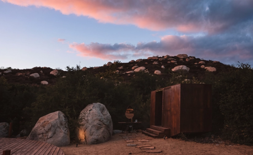 Sunset over a rocky hillside with wooden structures and a small fire pit.