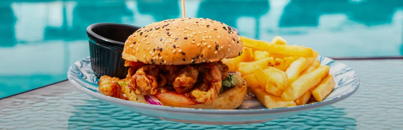 Burger with fries in a plate by a pool, bun covered with sesame seeds