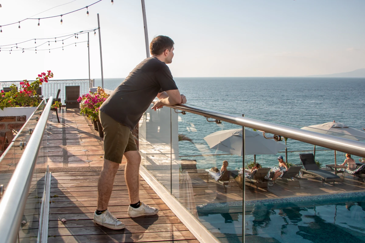 Man leaning on balcony railing by the ocean with sun loungers below.