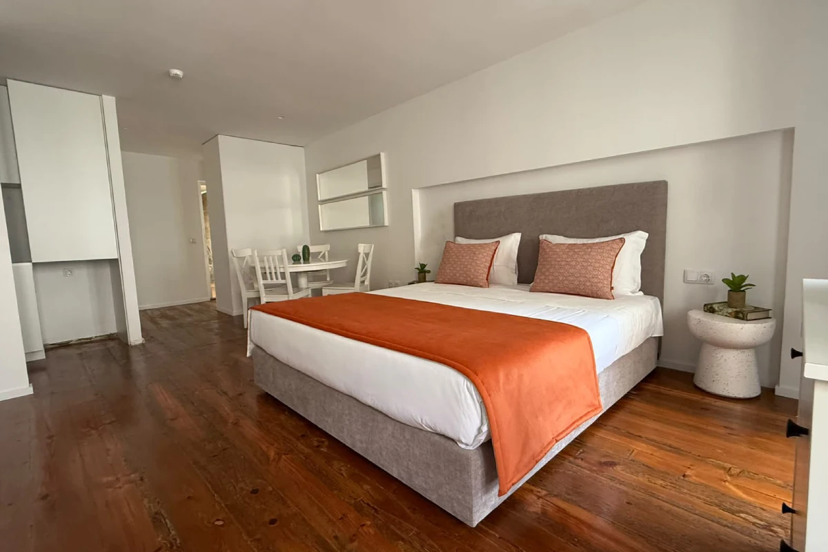 Dormitorio moderno con cama, cojines y mantas naranjas, luz natural y muebles blancos.