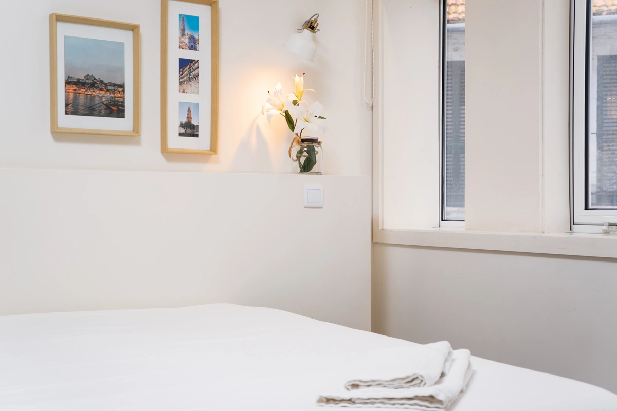 Dormitorio luminoso con cama blanca, flores en jarr&oacute;n y cuadros junto a ventanas