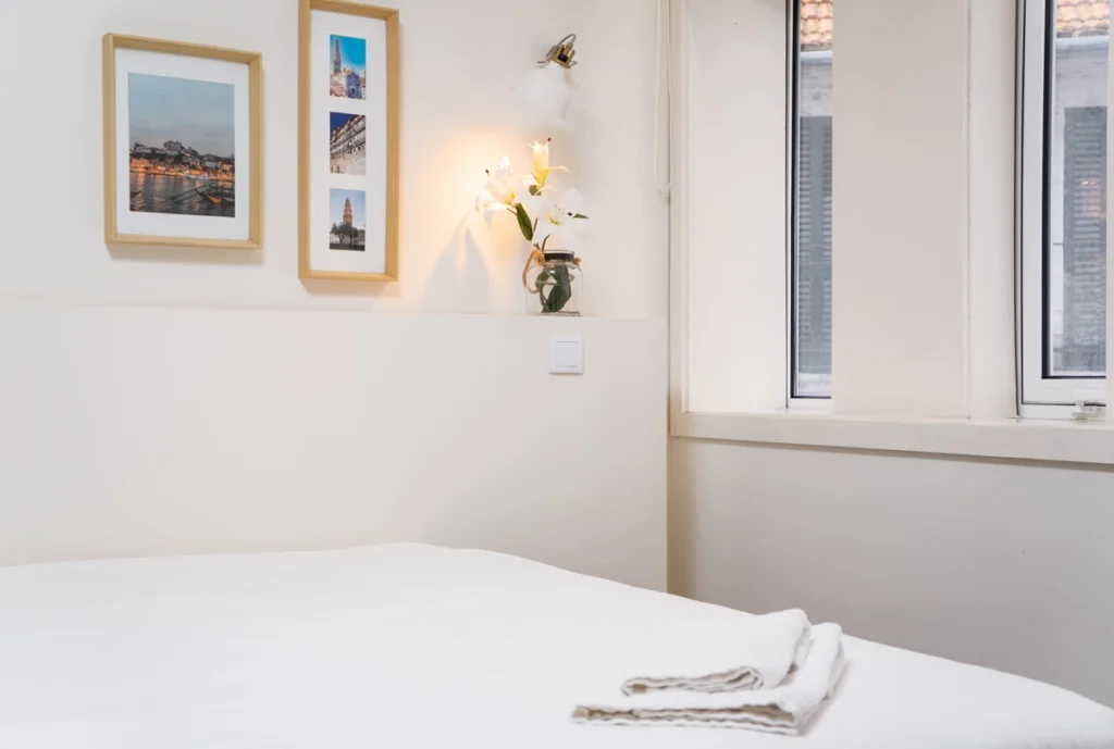 Dormitorio blanco con cama, cuadros y florero con lirios junto a la ventana