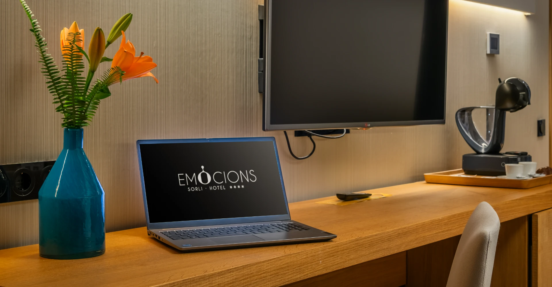 Televisor y port&aacute;til con flores naranjas y botella azul en una recepci&oacute;n de hotel
