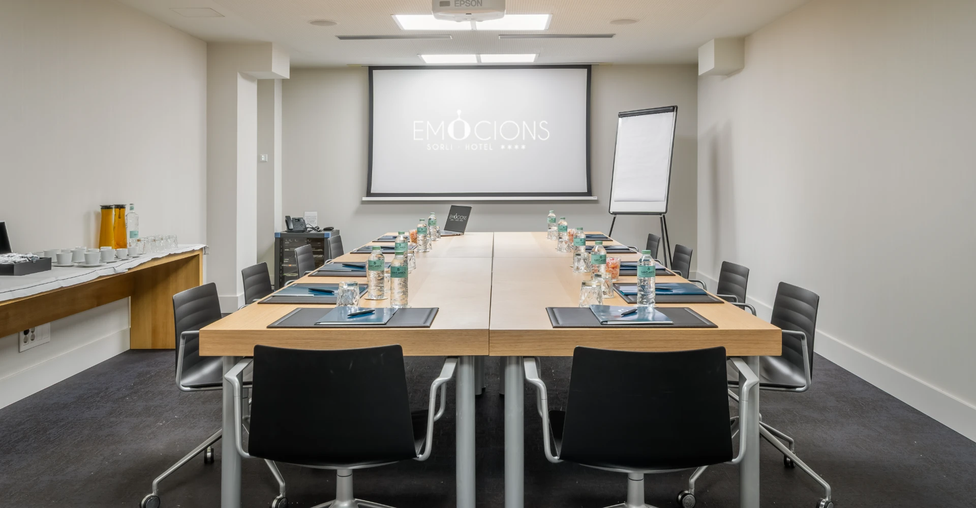 Sala de reuniones de hotel con mesa, sillas y pantalla de proyecci&oacute;n al fondo