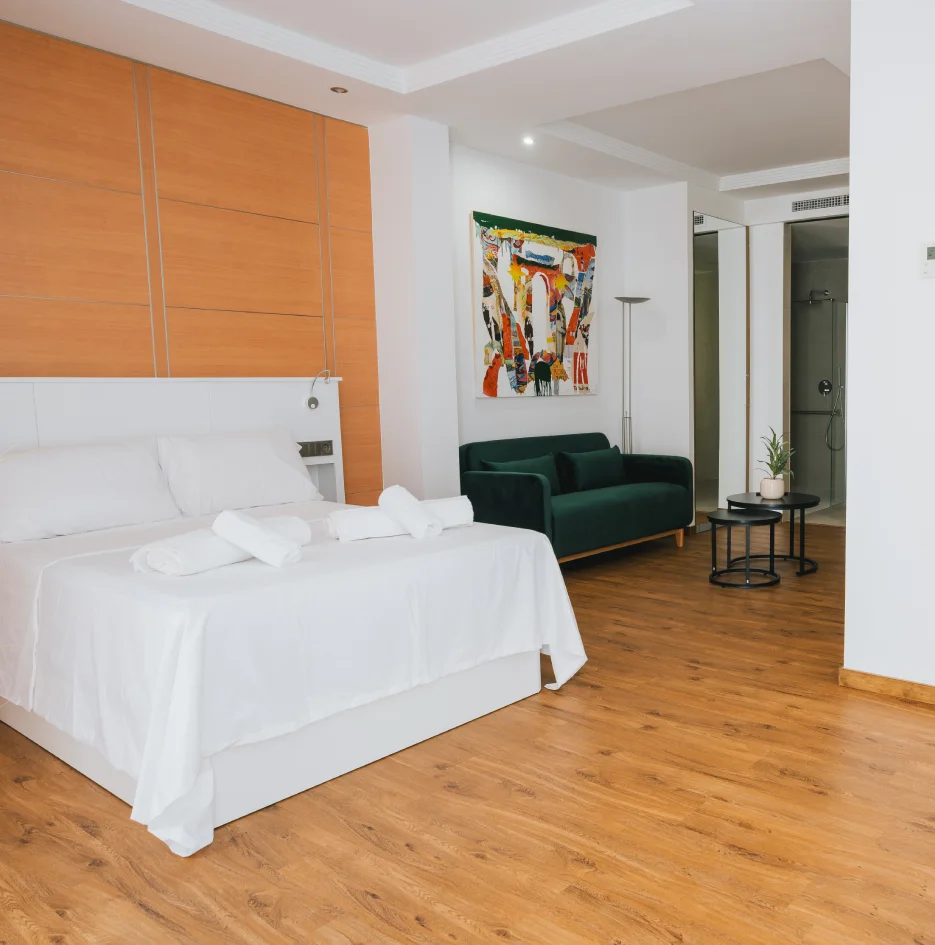 Habitaci&oacute;n hotel moderna con cama blanca y sill&oacute;n verde, vista a ba&ntilde;o cerrado