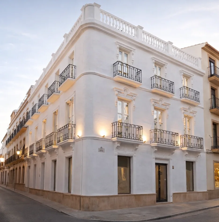 Fachada blanca de hotel con balcones de hierro y luces al atardecer.