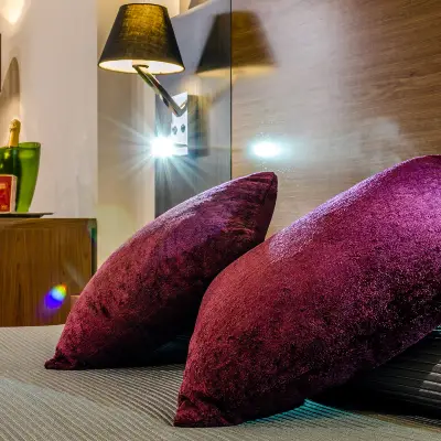 Dos cojines morados sobre cama, con l&aacute;mpara encendida al fondo
