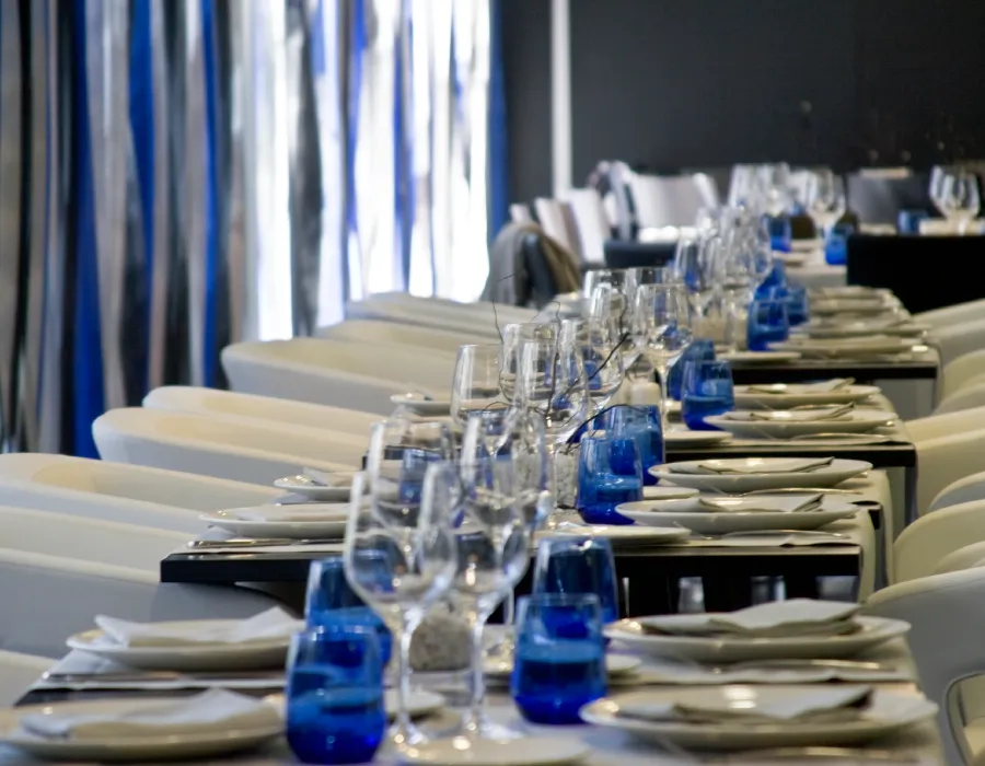Mesas de banquete con copas y vasos azules en un sal&oacute;n con cortinas.