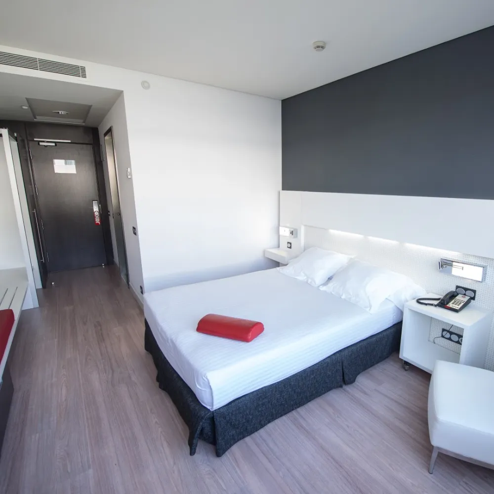 Habitaci&oacute;n de hotel moderna con cama blanca y puerta de acceso.