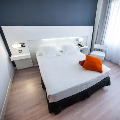 Habitaci&oacute;n moderna con cama blanca y coj&iacute;n naranja, escritorio y l&aacute;mparas