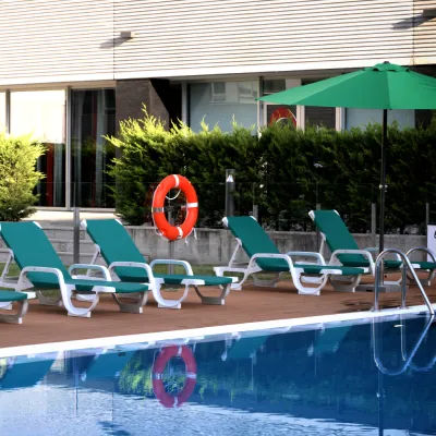 Piscina con sillas verdes y sombrilla, fondo de jard&iacute;n y edificio.