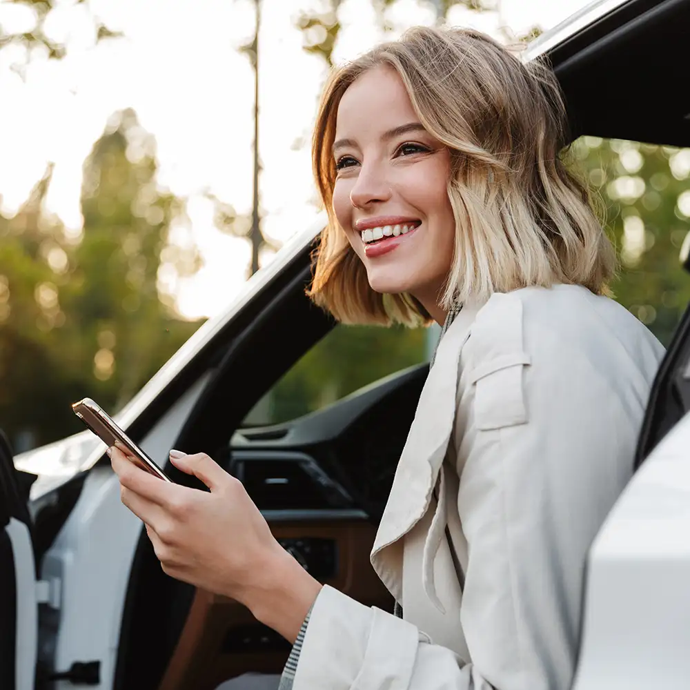 Mujer sonriente con tel&eacute;fono junto a un coche, al aire libre con luz de fondo