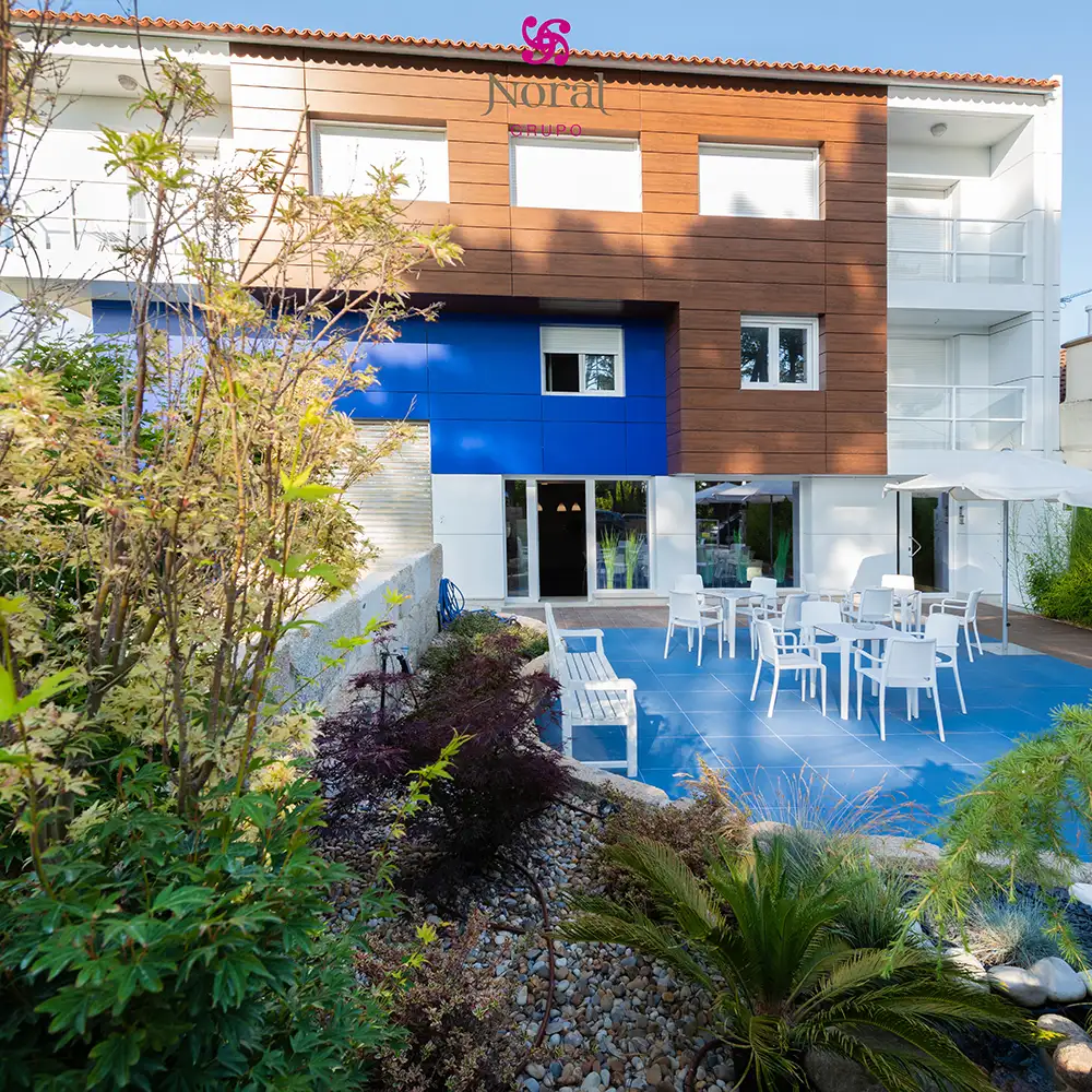 Fachada moderna con jard&iacute;n y terraza azul, mesas blancas y sillas.