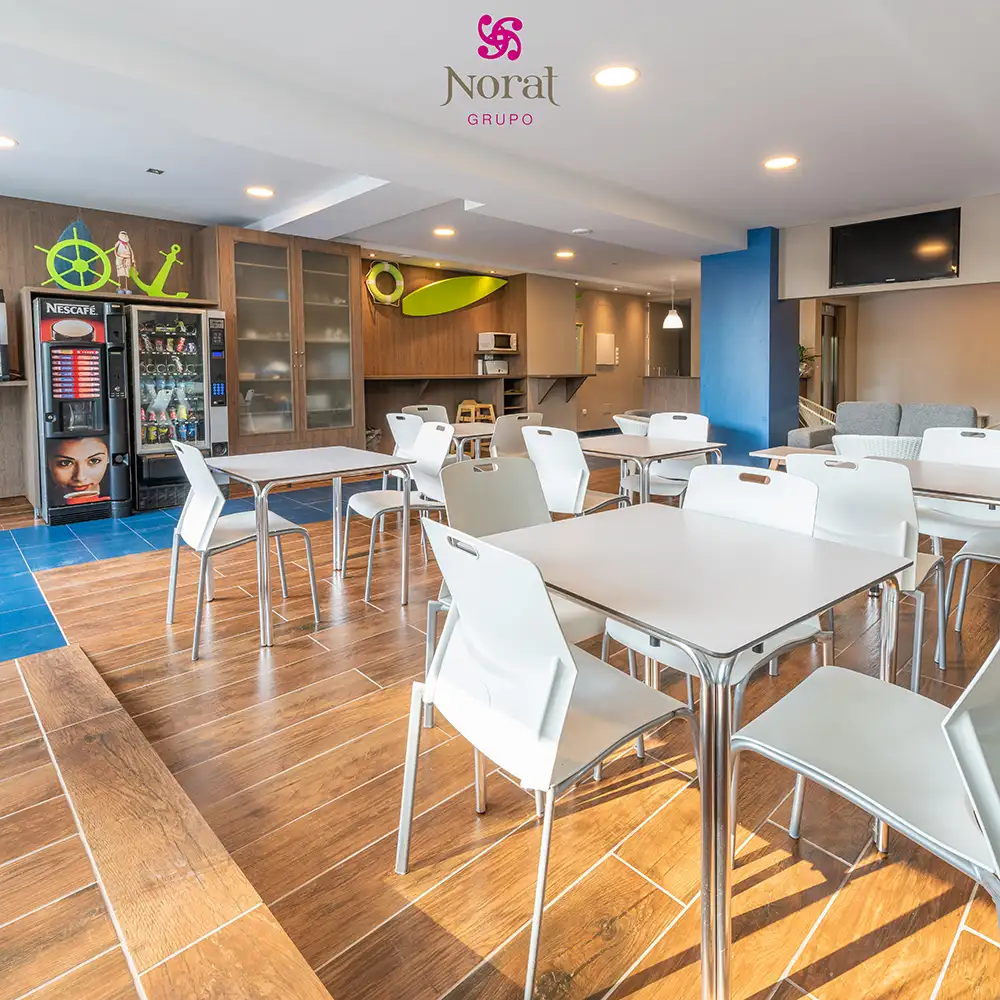 Sala de cafeter&iacute;a con mesas blancas y m&aacute;quina Nescaf&eacute;, logo Norat Grupo