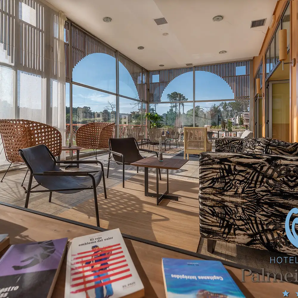Terraza interior con sillones, mesa y cortinas de vidrio; luz del d&iacute;a y cielo.