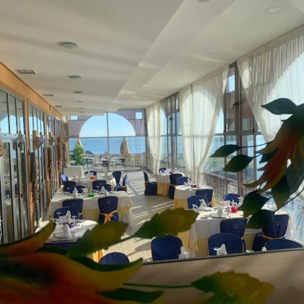 Sal&oacute;n de eventos con mesas y vista al mar, cortinas blancas y sillas azules