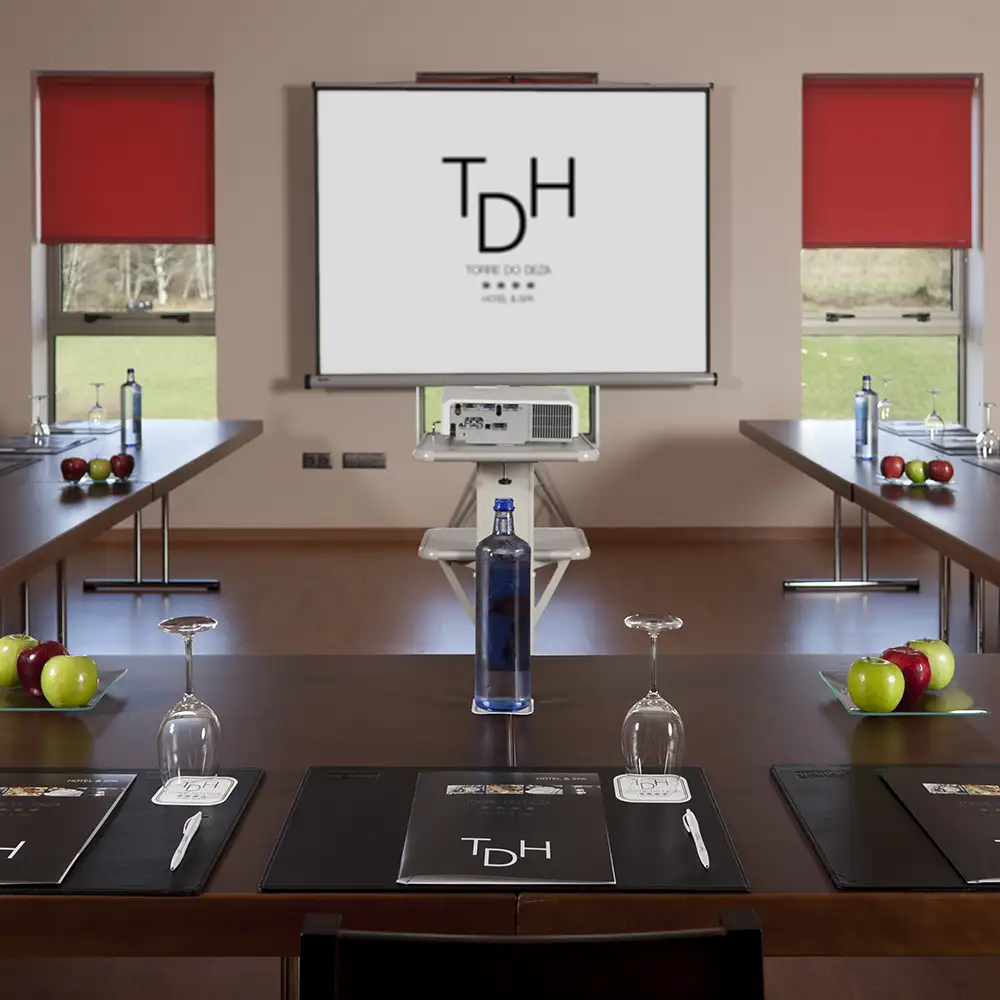 Sala de reuniones con proyector, mesas y persianas rojas abiertas y bebidas.
