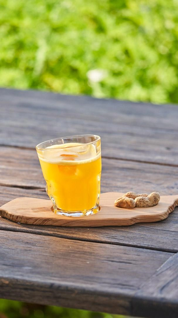 Copo com bebida amarela em mesa de madeira ao ar livre, com amendoins