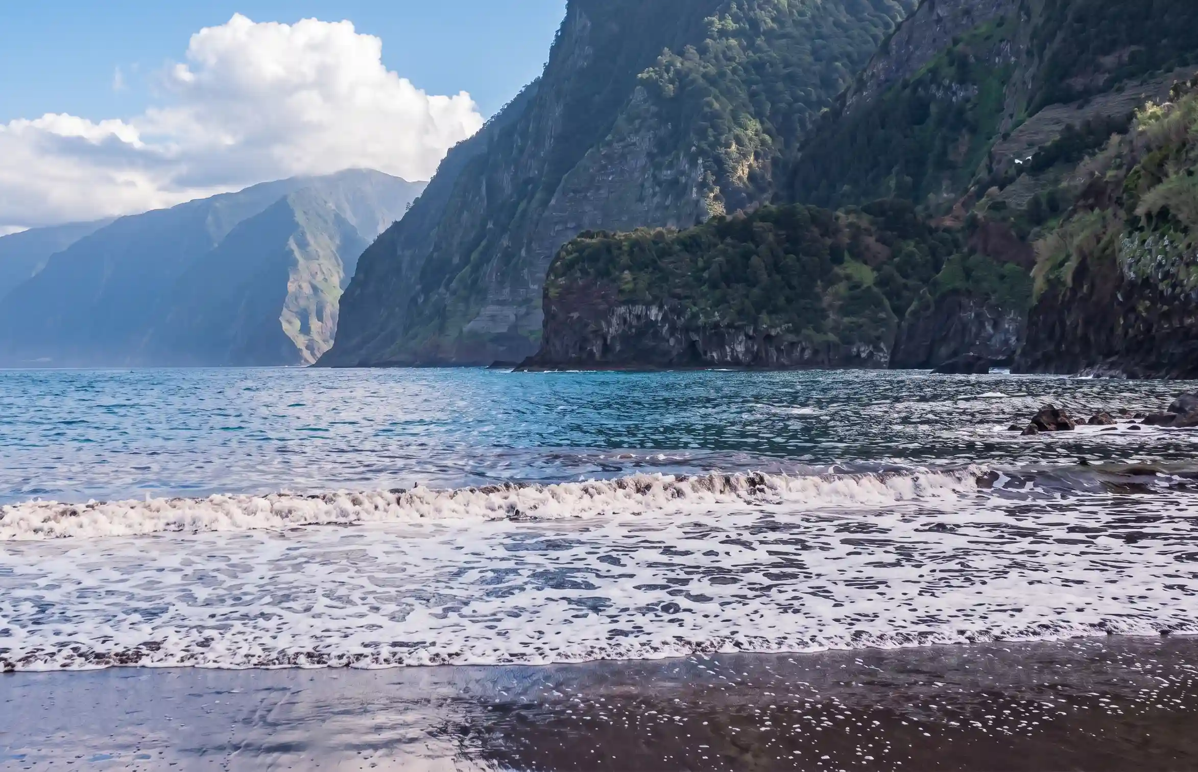 Praia com ondas e fal&eacute;sias verdes, montanhas ao fundo e c&eacute;u nublado