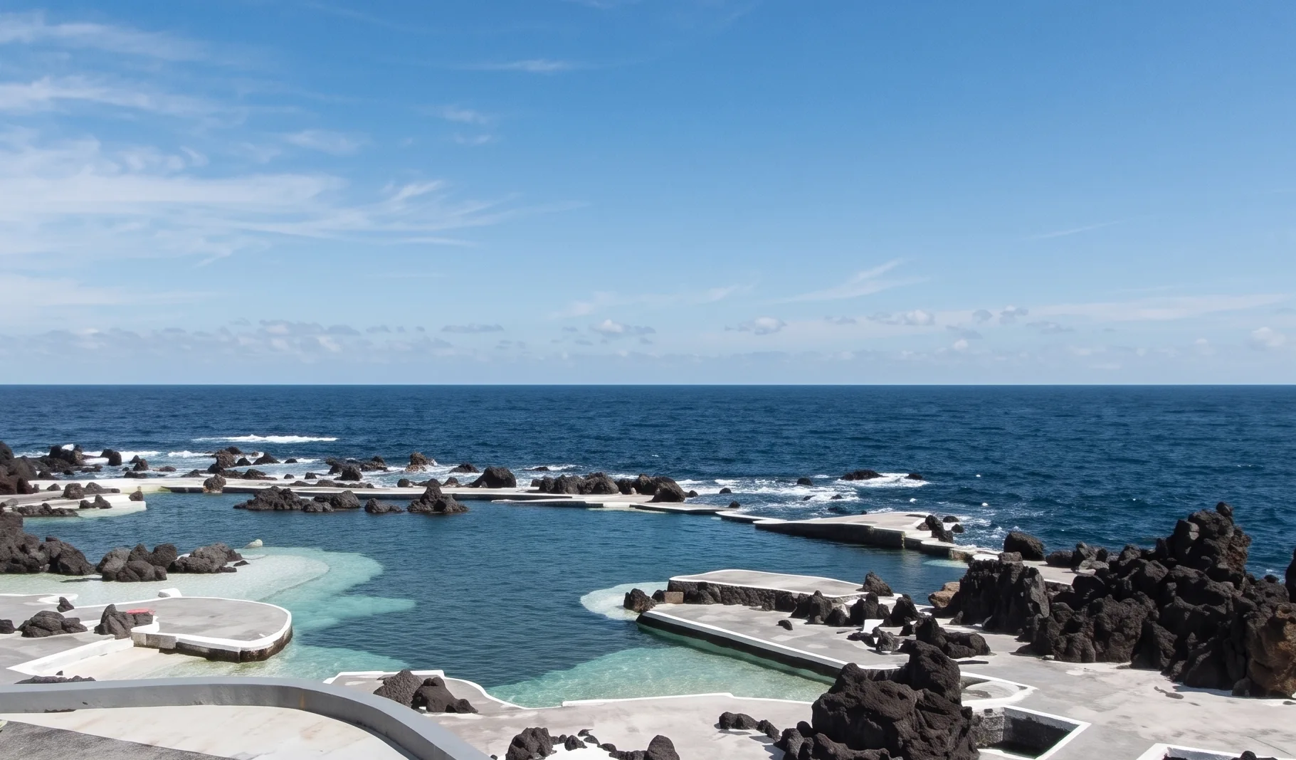 Piscina de borda branca com rochas vulc&acirc;nicas e mar ao fundo sob c&eacute;u azul