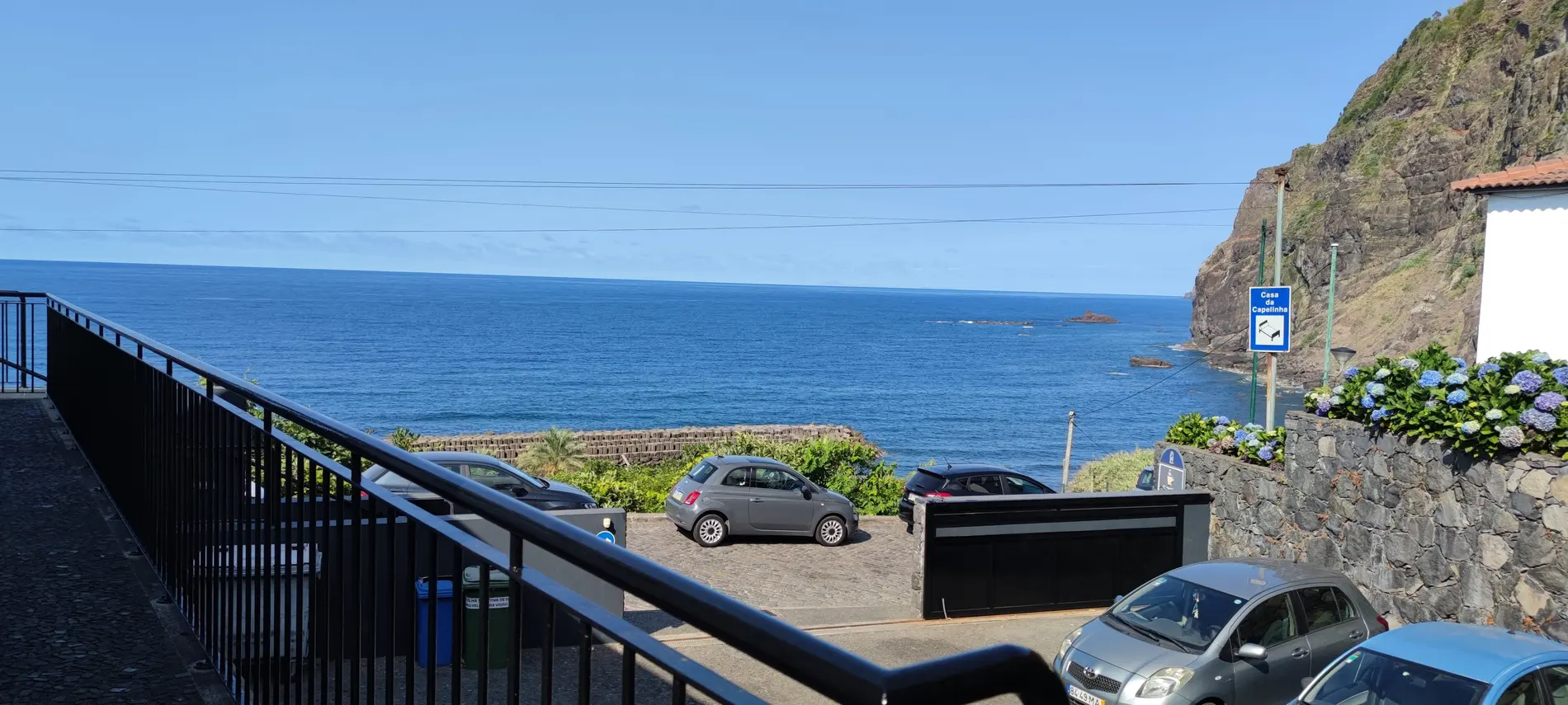 Vista do oceano e entrada com carros, placa &ldquo;Casa da Capelinha&rdquo; e rocha costeira
