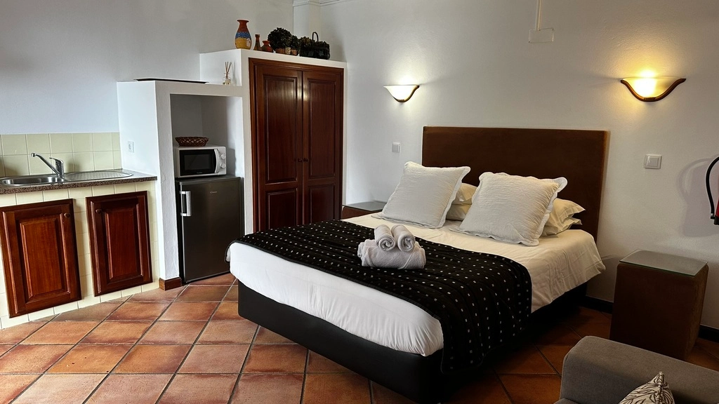 Quarto de hotel com cama, travesseiros, cozinha compacta e ilumina&ccedil;&atilde;o quente
