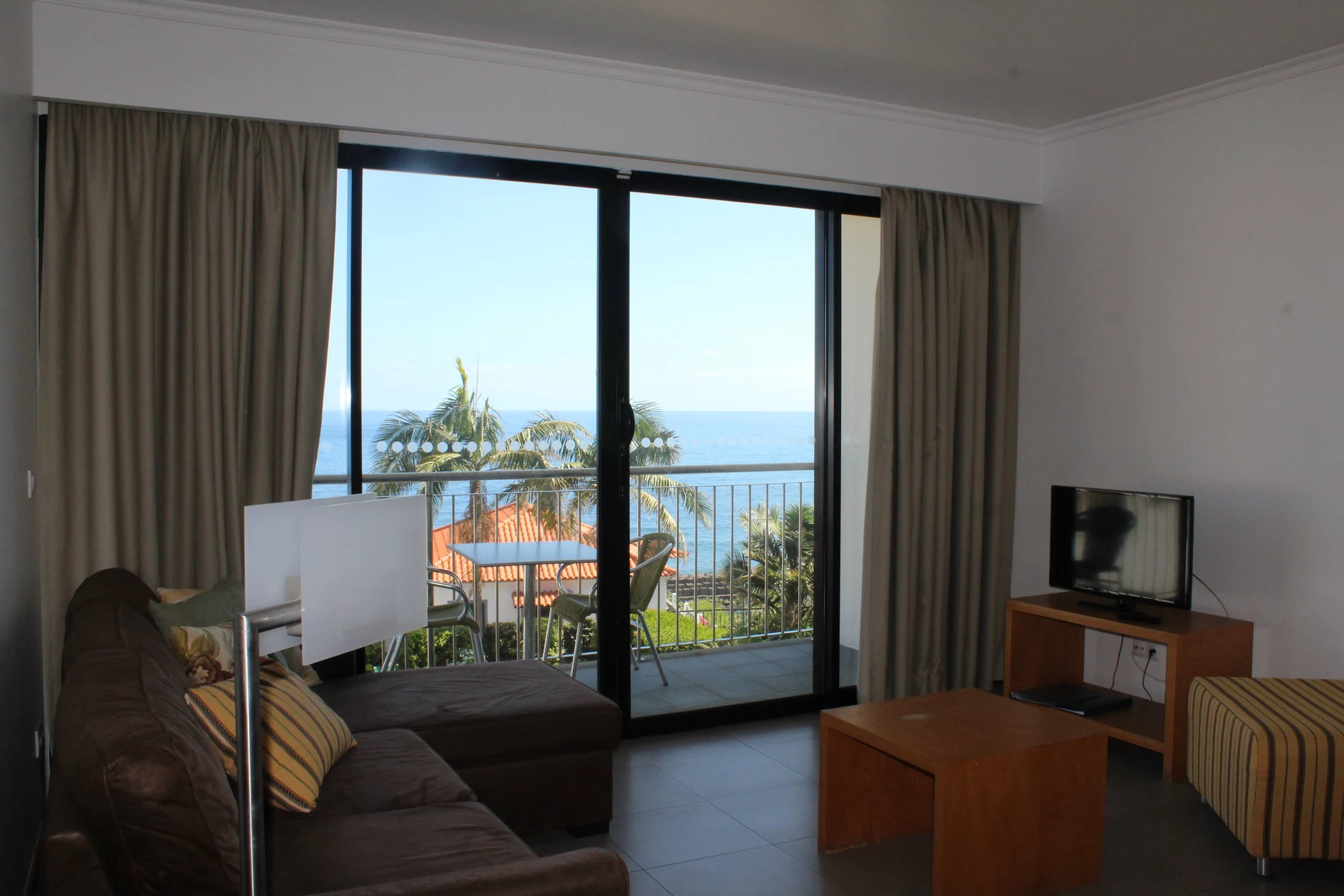 Sala com cortinas e vista para o mar, varanda e palmeiras ao fundo
