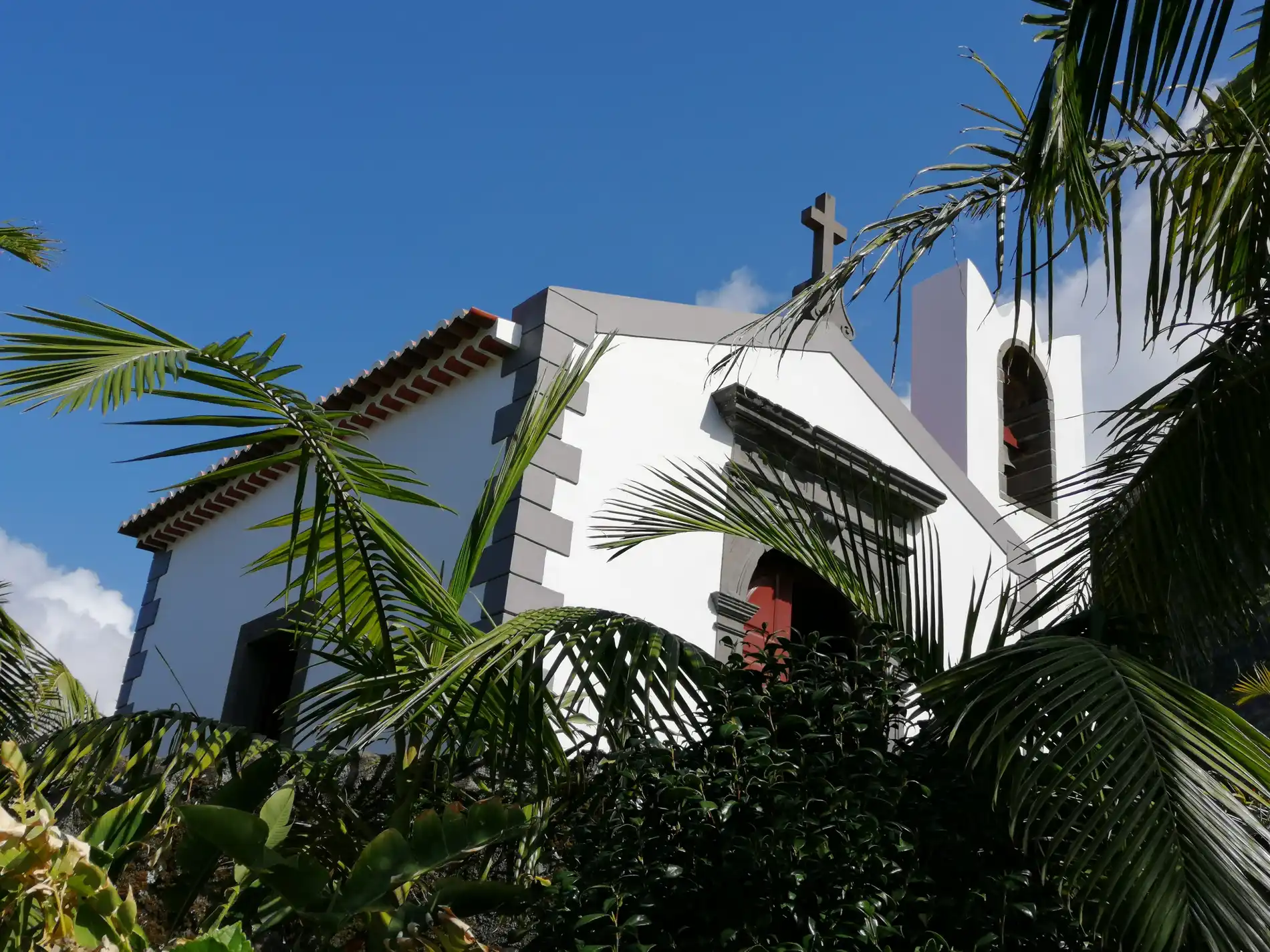 Igreja branca com cruz e palmeiras ao c&eacute;u azul.