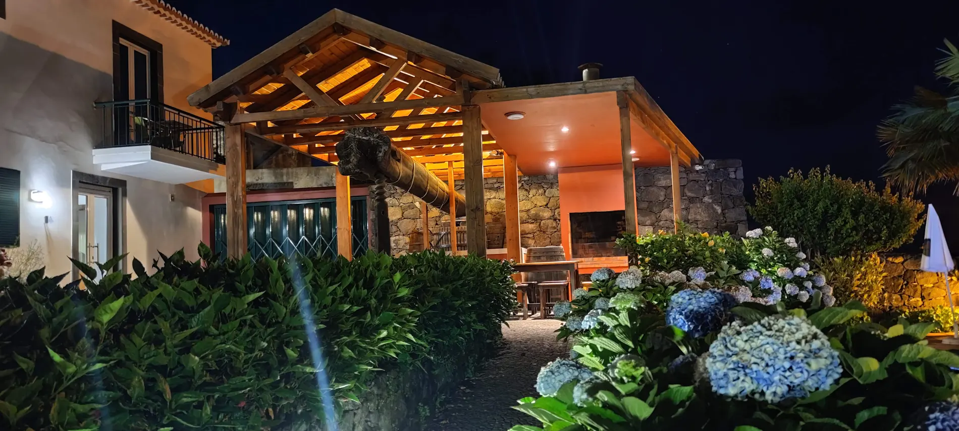 Varanda de madeira iluminada &agrave; noite, com jardim de flores e pedra ao fundo