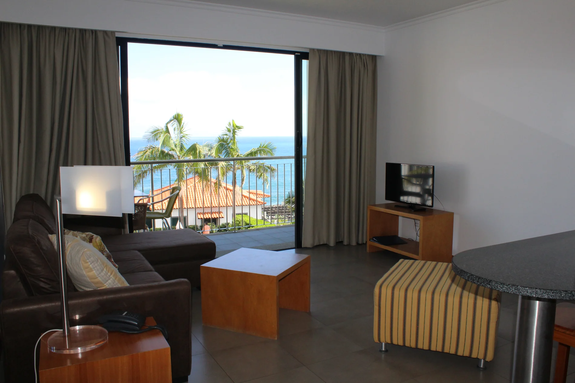 Sala de hotel com vista para o oceano e palmeiras atrav&eacute;s de janela ampla