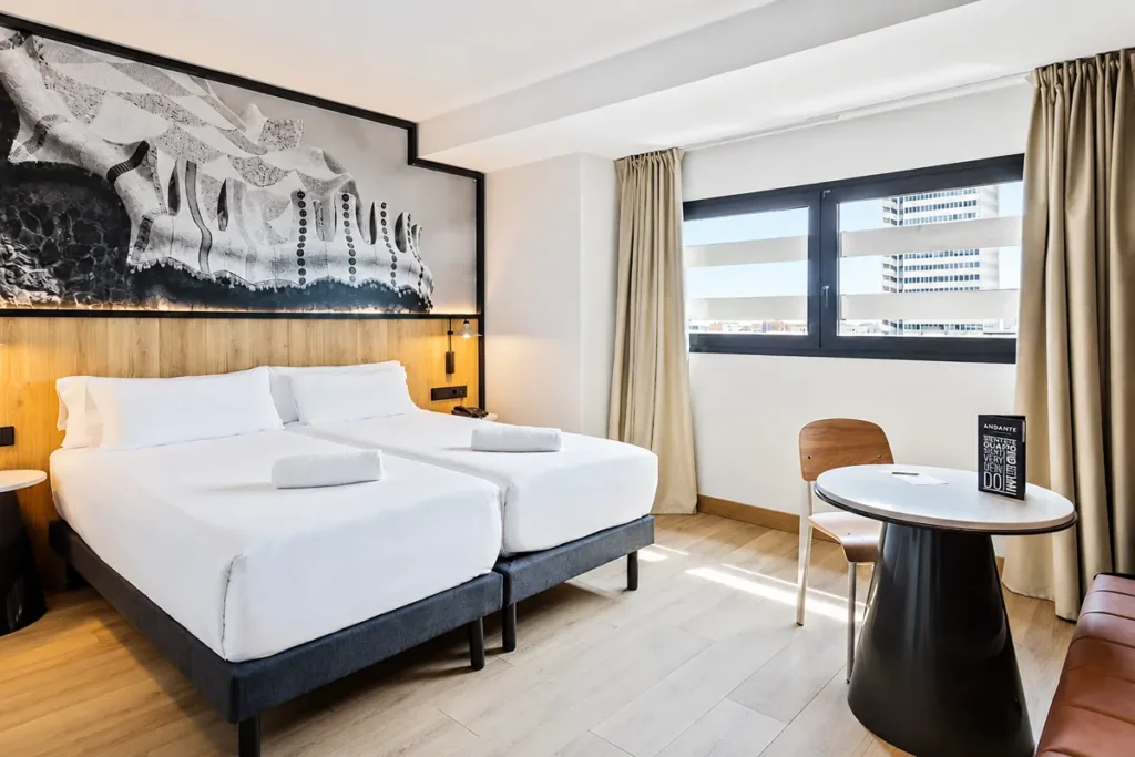 Habitaci&oacute;n de hotel moderna con cama doble, mesa redonda y grandes ventanales